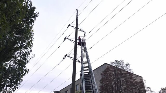An electrician repairs a line of high-voltage wires. Bottom view of the retractable ladder and the cradle in which the worker stands. Trees, sky and building are visible in the background