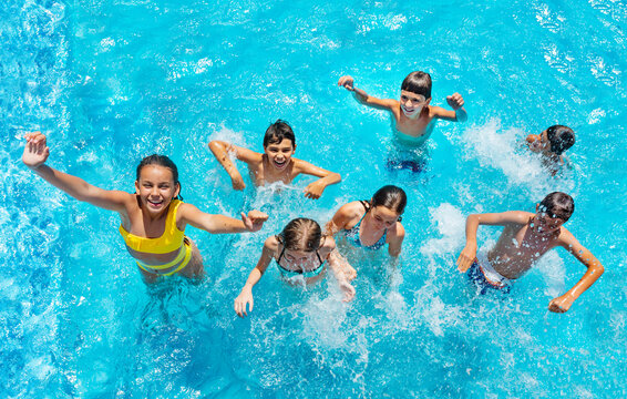 Group of many kids jump and play in swimming pool, splashing, lift hands smile view from above