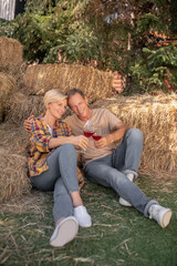 Naklejka na ściany i meble Middle-aged couple sitting on hay, toasting with red wine