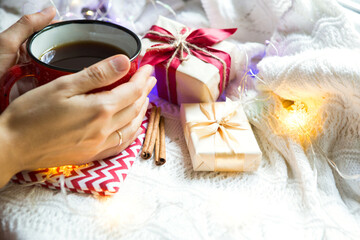 Fototapeta na wymiar A woman's hand in a warm sweater holds a red mug with a hot drink on a table with Christmas decorations. New year's atmosphere, cinnamon sticks and a slice of dried orange, gifts, garland and tinsel