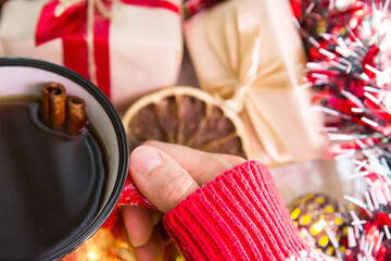 Obraz na płótnie Canvas A woman's hand in a warm sweater holds a red mug with a hot drink on a table with Christmas decorations. New year's atmosphere, cinnamon sticks and a slice of dried orange, gifts, garland and tinsel