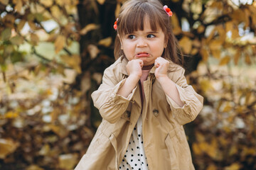 Beautiful little girl in a beige coat shows angry in the autumn park