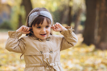 Happy and beautiful girl in a beige coat shows emotions and closes her ears with her fingers in the autumn park