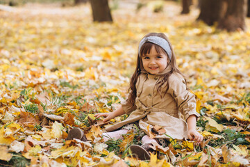 Happy and beautiful little girl in a beige coat sitting among the yellow leaves in the autumn park