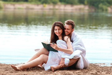 caucasian couple wrapped in a blanket lie on the riverbank and read a book. Self isolation from people. Unity with nature and digital detox