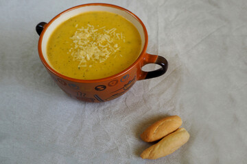 a soup of zucchini, onions,potatoes and carrots, sprinkled with grated hard cheese, in a brown plate with handles stands on a gray tablecloth with two small Italian loaves side view . home cooking
