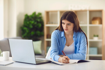 Young female student writing in notebook while watching online lecture sitting at desk at home
