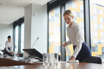 Portrait of smiling young woman cleaning table with sanitizing spray in conference room while...