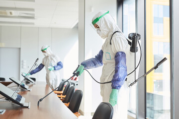 Side view portrait of two workers wearing hazmat suits disinfecting conference room in office, copy space