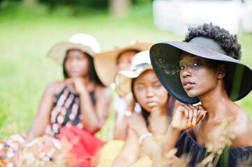 Group of four gorgeous african american womans wear summer hat sitting at green grass in park.