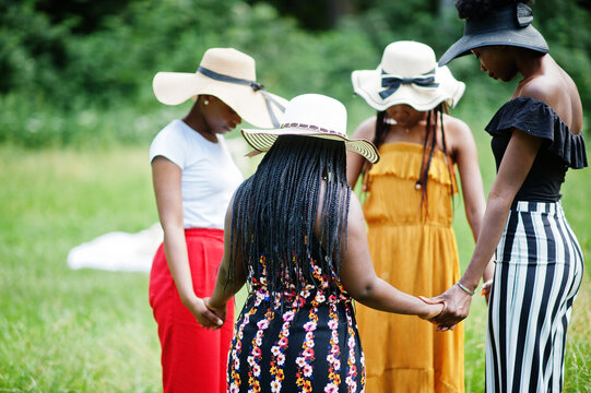 Group of four gorgeous african american womans wear summer hat holding hands and praying at green grass in park.