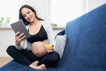 Smiling pregnant brunettre woman with a glass of orange juice is sitting on a sofa in the living room and reading articles on tablet. She is wearing sports clothes.