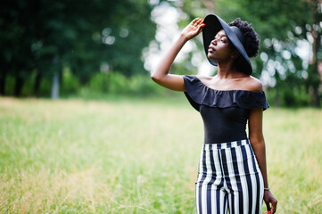 Obraz premium Portrait of gorgeous african american woman 20s in wear in black and white stripe pants and summer hat posing at green grass in park.