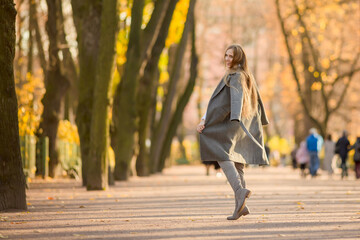 Happy young woman walks in the autumn park. An attractive woman with light brown hair and a gray coat is walking down the alley. Beautiful autumn mood. Fall season.