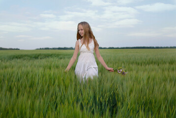 Slender blonde girl in a white sundress walks on a green field