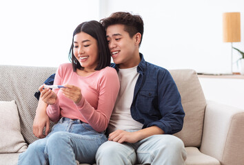 Joyful Asian Couple Holding Positive Pregnancy Test Sitting At Home