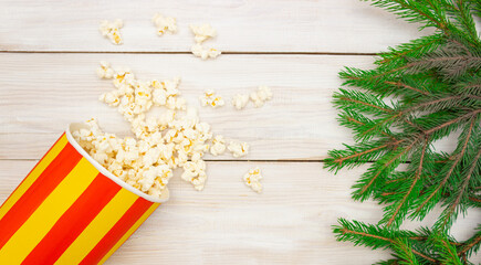 The concept of cinema, New Year and Christmas. Popcorn spills out of the box, green Christmas tree branches. Light wooden background, free space for text, top view.