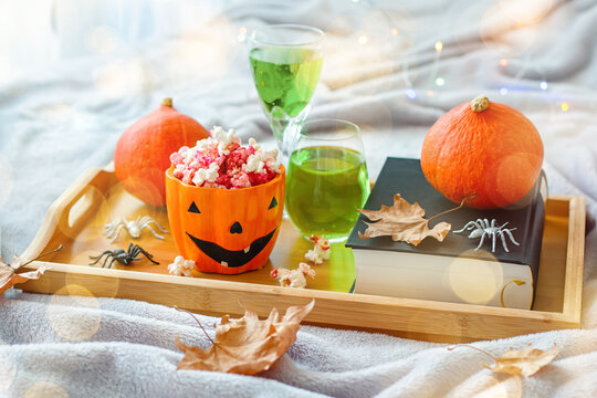 Book, halloween decor, candles, popcorn and drinks on tray on bed, halloween reading concept with bokeh background. Still life for Halloween at home