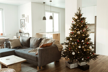 Minimalist modern home interior design concept. Comfortable cozy living room decorated with Christmas tree with gifts, sofa, plaid. Christmas / New Year celebration decorations.