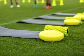 Naklejka premium Training mat, goal roller and balance cushion. Sports training equipment on grass field. Young athletes and training cones in blurred background