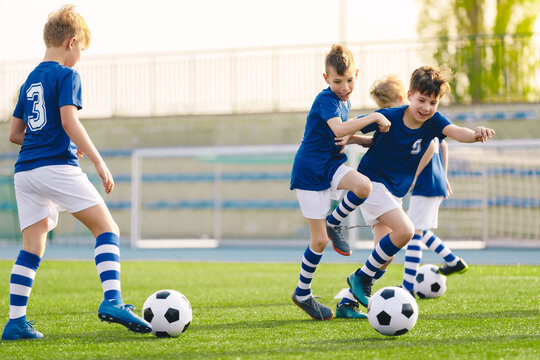 Young boys playing football training game. Happy children kicking soccer balls on practice pitch. Soccer training stadium in the background. School age boys in soccer blue jersey shirts