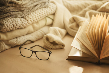 Soft, warm sweaters are stacked on the bed, along with an open book and black-rimmed glasses in the sunlight. Home comfort. Reading process.