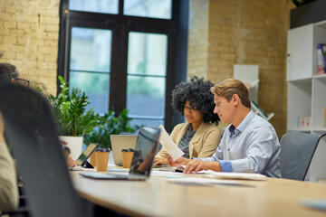 Two male and female coworkers looking at laptop screen and analyzing project results while sitting together at the table in the modern office or coworking space