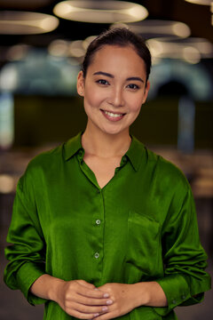 Vertical Shot Of A Young Happy Asian Business Woman In Green Silk Shirt Looking At Camera And Smiling While Posing In The Office