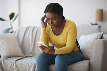 Worried black lady looking at smartphone screen, sitting on couch at home