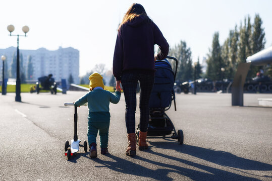 Mom With Stroller And Baby With Scooter Walking Together In Park, Portrait From Back, Parental Care, Happy Childhood