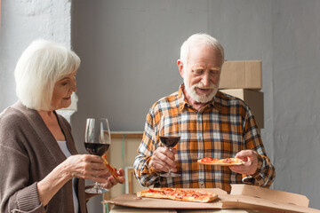 senior couple holding pieces of pizza and glasses of wine in new house