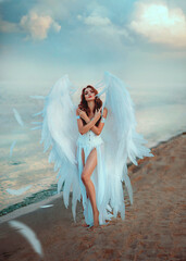 Young beautiful woman fallen angel stands on the sea beach enjoy nature. Creative sexy costume, huge artificial bird wings and white vintage dress with slits. Adult girl with seductive long legs