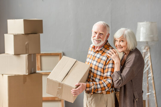 senior man holding cardboard box and wife leaning on shoulder in new house, moving concept