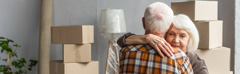panoramic shot of senior woman with closed eyes hugging husband in new house, moving concept