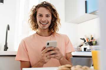 Caucasian handsome smiling guy using cellphone while having breakfast