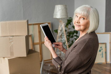 senior woman holding digital tablet with blank screen and looking back with cardboard boxes on background, moving concept
