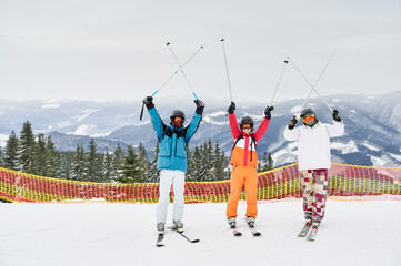 Three cheerful tourists posing on snowy slope against mountainous background, holding their ski poles up above. Friends at high spirits having fun at ski resort. Concept of winter sport and friendship