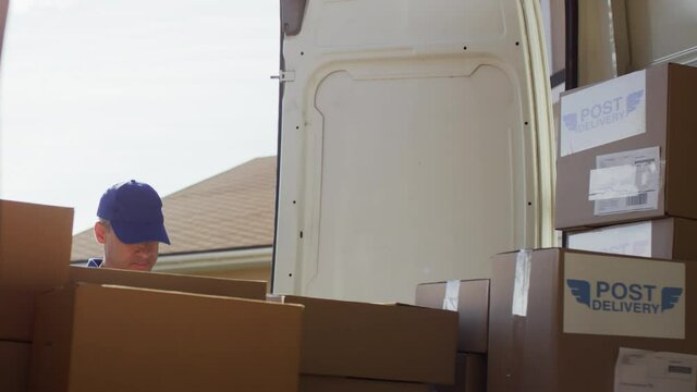 Handheld shot from inside of mail van: serious male courier in blue uniform taking package out of trunk