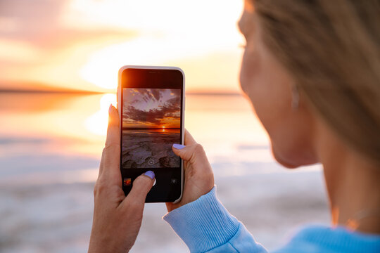 Young happy blonde woman taking photo of sunset on cellphone outdoors