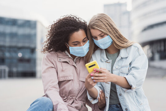 Young women friends wering protective face masks using smartphone in a city, Cute smiling girls having fun on a city street, Technology, people, healthy lifestyle, self care, friendship concept - Powered by Adobe