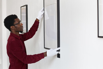 Side view at African-American man hanging blank frames on wall while planning art gallery or exhibition, copy space