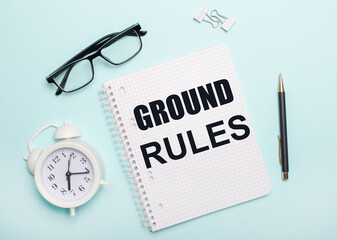 On a light blue background lie black glasses and a pen, a white alarm clock, white paper clips and a notebook with the words RULES OF THE EARTH. Business concept