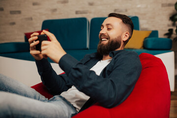 Smiling bearded man sit on the red armchair in the living room and relax at home with smartphone in hands.