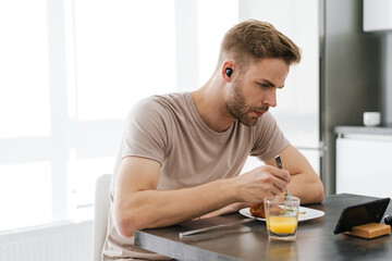 Fototapeta na wymiar Young man in earphones looking at cellphone while having breakfast at home