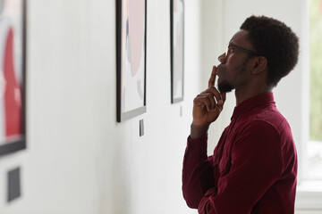Fototapeta na wymiar Side view portrait of young African-American man looking at paintings and thinking at art gallery or museum exhibition, copy space