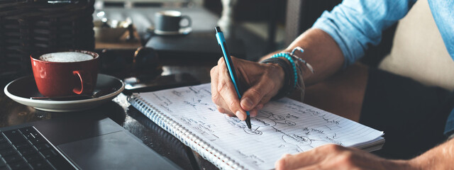 Entrepreneur working on his business project in city cafe, writing down ideas using pen and paper, close-up. Wide screen, panoramic