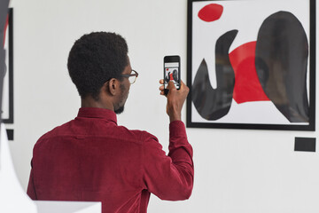 Back view portrait of young African-American man taking photo of painting via smartphone at...