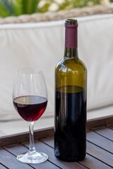 One glass of red wine and an open dark green glass bottle on a wooden serving table near a beige sofa