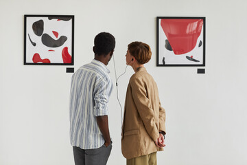 Graphic back view portrait of two young people looking at paintings and sharing audio guide while...