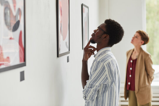 Side view portrait of young African-American man looking at paintings while exploring modern art gallery exhibition, copy space